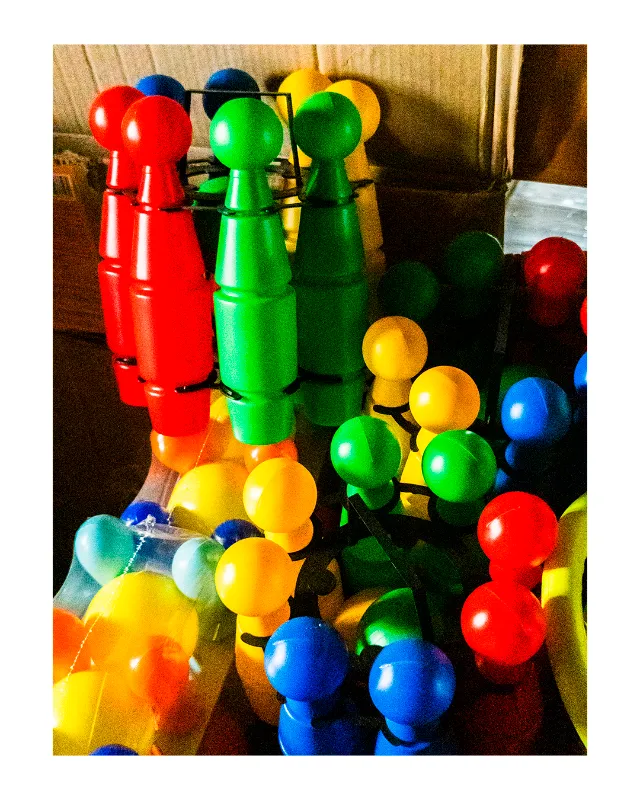 Collection de quilles et boules en plastique brillant rouge, vert, jaune et bleu disposées sur une table, éclairage naturel latéral créant des reflets lumineux, ambiance nostalgique des jeux vintage