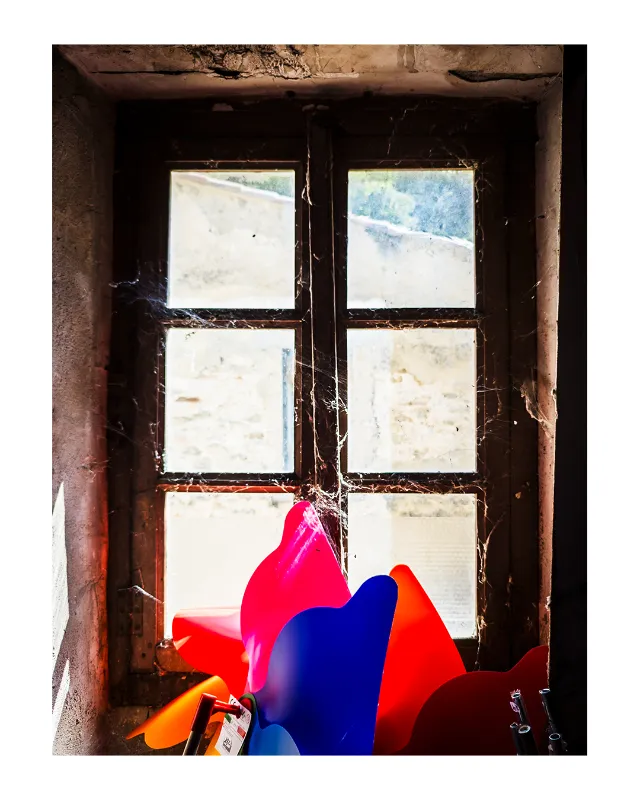 Jouets roses, bleus et oranges vibrants disposés devant une fenêtre à six carreaux patinée par le temps, dans l'embrasure d'un mur ancien aux tons ocre et bruns, créant un contraste saisissant entre modernité colorée et architecture abandonnée