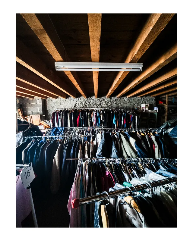 Perspective intérieure d'un magasin de vêtements aux Grands Magasins Universelles de Dieulefit, Drôme, avec poutres en bois brut apparentes créant des lignes géométriques, mur en pierre, portants de vêtements colorés et éclairage industriel