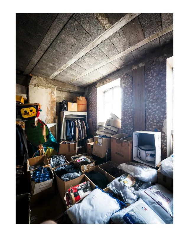 Photographie d'intérieur d'un magasin vintage à Dieulefit dans la Drôme, montrant un espace de stockage avec plafond en béton brut, murs patinés, cartons de vêtements au sol, portants et lumière naturelle latérale créant un contraste dramatique