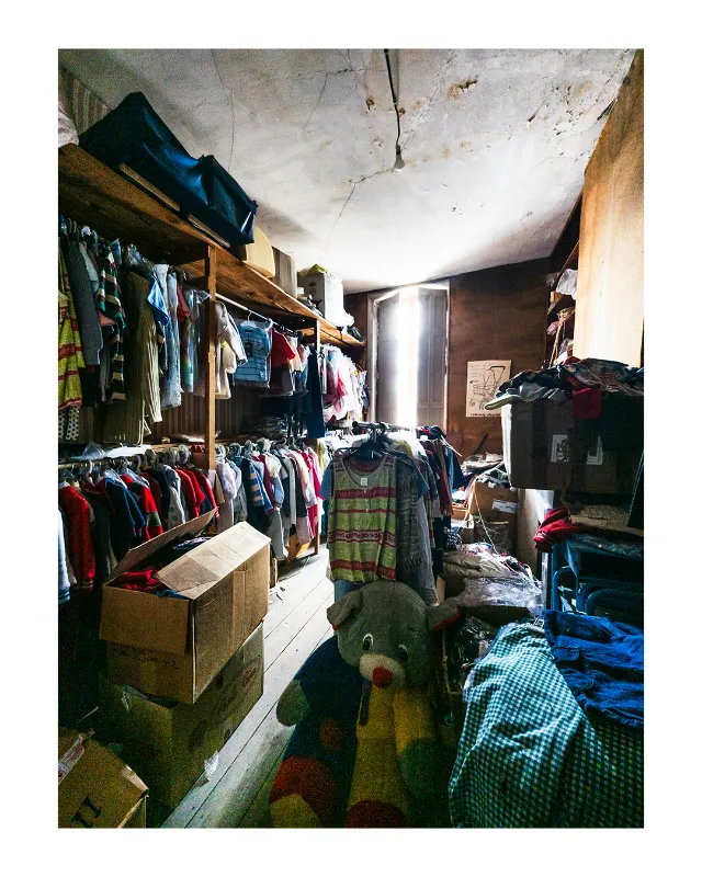 Photographie documentaire d'un magasin vintage à Dieulefit : étagères en bois débordant de vêtements multicolores, cartons au sol, plafond en béton brut, lumière naturelle latérale créant une atmosphère authentique et nostalgique