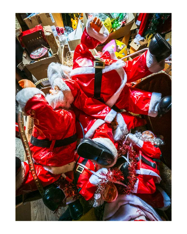 Vue en plongée sur un Père Noël en costume rouge et blanc assis au milieu d'un bazar de cadeaux emballés, décorations de Noël et cartons dans un magasin de jouets à Dieulefit
