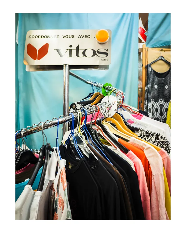 Portant métallique garni de vêtements multicolores sur cintres, dominé par une enseigne vintage Vitos Paris sur fond de rideaux bleu turquoise, photographié dans les grands magasins universels de Dieulefit