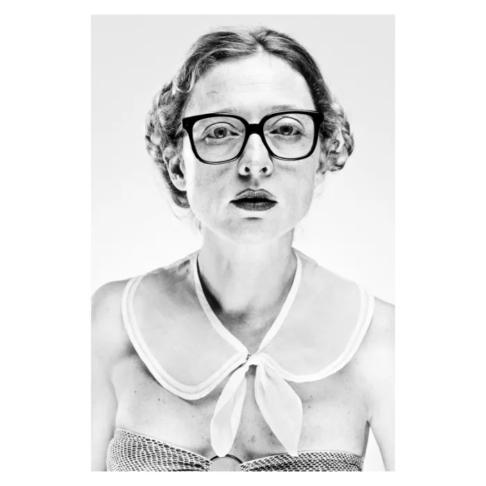 Portrait artistique féminin en noir et blanc au style rétro, femme portant des lunettes à monture noire épaisse et un col Claudine blanc, regard frontal sérieux, photographie de studio réalisée à Bagnolet par Nicolas Louis.