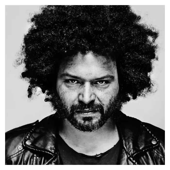 Portrait artistique en noir et blanc d'un homme au regard frontal intense et barbe sombre, arborant une coupe afro volumineuse et un blouson en cuir, photographie de studio réalisée à Bagnolet par Nicolas Louis.