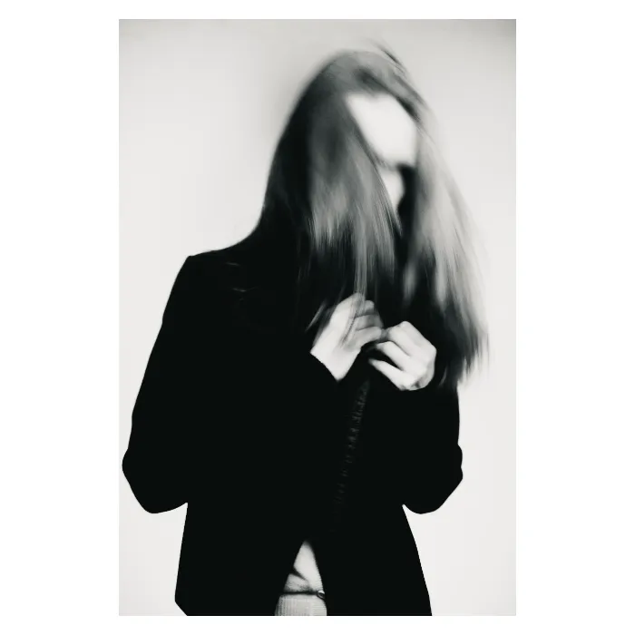 Portrait artistique en noir et blanc avec un effet de flou de bougé dynamique, silhouette d'une femme aux longs cheveux voilant son visage, mains ajustant le col d'une veste sombre, atmosphère onirique et abstraite, photographie de studio réalisée à Bagnolet par Nicolas Louis.