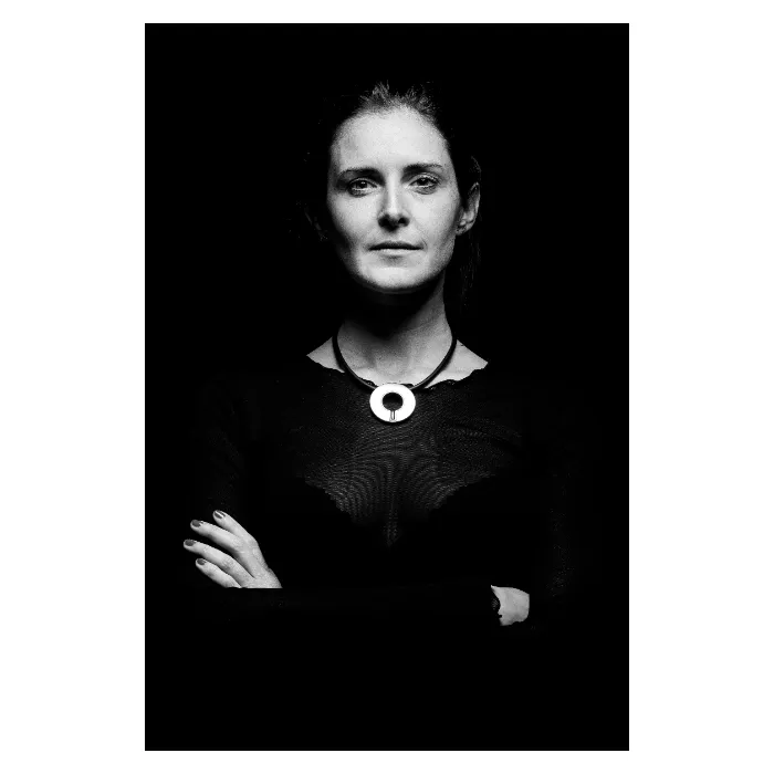 Portrait frontal en noir et blanc d'une femme au regard déterminé, bras croisés sur la poitrine, portant un vêtement noir texturé et un collier imposant avec un pendentif circulaire argenté, photographie de studio sur fond noir profond réalisée à Bagnolet par Nicolas Louis.