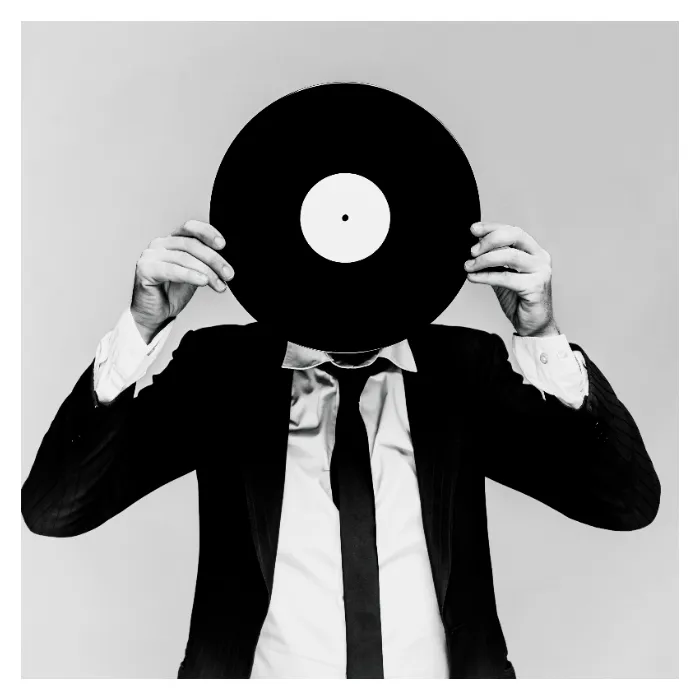 Portrait conceptuel en noir et blanc de l'artiste Daprinsky sur fond gris clair, le visage entièrement masqué par un disque vinyle tenu à deux mains, portant un costume sombre et une cravate fine, photographie de studio graphique réalisée à Bagnolet par Nicolas Louis.