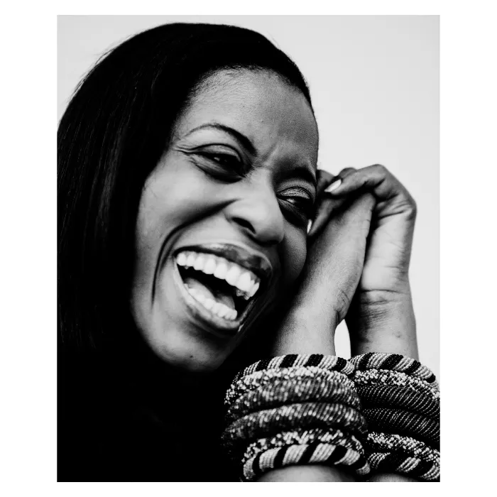 Portrait en noir et blanc très serré d'une femme riant aux éclats, expression de joie spontanée, mains jointes près du visage et accumulation de bracelets tressés au poignet, photographie de studio contrastée réalisée à Clichy par Nicolas Louis.