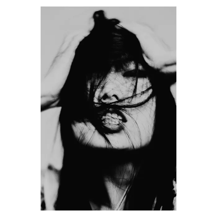 Portrait expressionniste en noir et blanc d'une femme aux longs cheveux noirs ébouriffés, les mains dans la chevelure et des mèches balayant son visage, pose dynamique et intense, photographie de studio contrastée réalisée au Vésinet par Nicolas Louis.