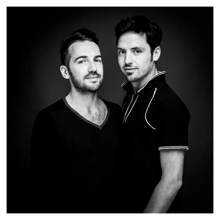 Portrait de complicité en noir et blanc des deux hommes, Tony et Régis, cadrés en plan serré sur fond sombre, l'un portant un pull à col en V et l'autre un polo noir à liserés blancs, photographie de studio contrastée réalisée à Bagnolet par Nicolas Louis.