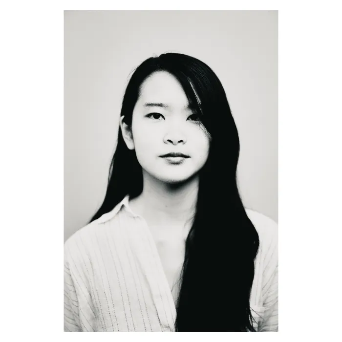Portrait frontal en noir et blanc d'une jeune femme au regard calme et déterminé, longs cheveux noirs lisses tombant sur une épaule, portant une chemise claire à fines rayures verticales, photographie de studio épurée réalisée au Vésinet par Nicolas Louis.