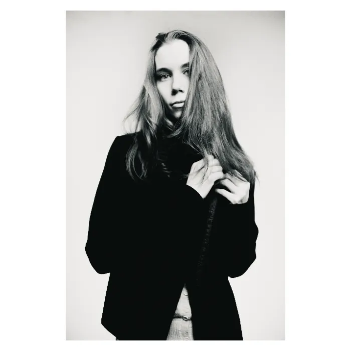 Portrait en noir et blanc d'une jeune femme aux longs cheveux blonds, une main ramenant une mèche sur son épaule, portant une veste sombre sur un gilet clair, expression sereine et pensive, photographie de studio épurée réalisée à Bagnolet par Nicolas Louis