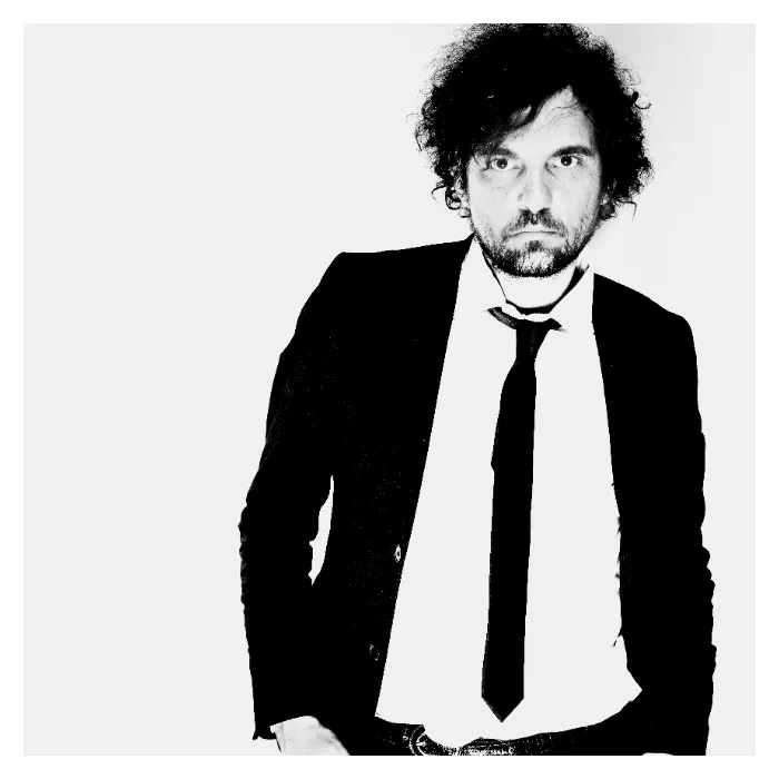 Portrait frontal en noir et blanc de l'artiste Daprinsky sur fond blanc, visage cadré de près avec un regard direct et intense, portant un costume sombre, une chemise blanche et une cravate fine noire, photographie de studio au contraste élevé réalisée à Bagnolet par Nicolas Louis.