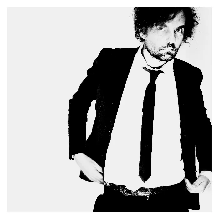 Portrait en noir et blanc de l'artiste Daprinsky sur fond blanc, portant un costume sombre ajusté, une chemise blanche et une cravate fine noire, attitude rock et regard intense, photographie de studio contrastée réalisée à Bagnolet par Nicolas Louis.