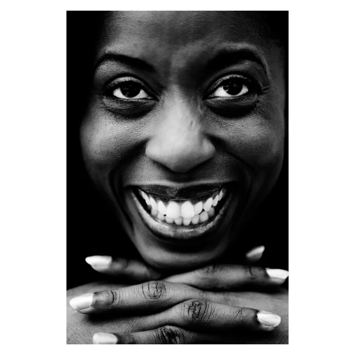 Portrait frontal en noir et blanc d'une femme au sourire éclatant, visage cadré de près avec les mains croisées sous le menton, éclairage contrasté mettant en valeur les détails du regard et de la peau, photographie de studio réalisée à Clichy par Nicolas Louis.