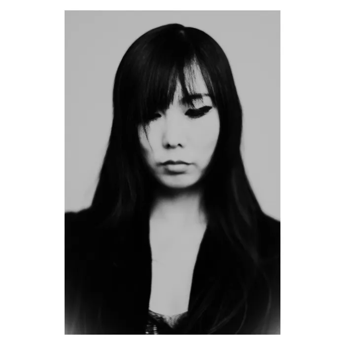 Portrait frontal en noir et blanc d'une femme aux longs cheveux sombres et frange, regard baissé dans une expression pensive et mélancolique, portant un vêtement sombre, photographie de studio épurée réalisée au Vésinet par Nicolas Louis.