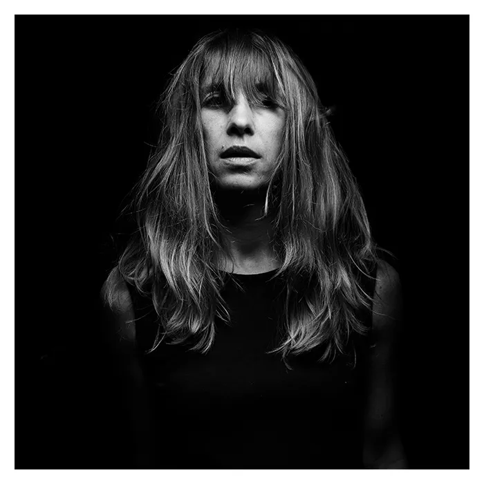Portrait frontal en noir et blanc d'une femme aux longs cheveux blonds vénitiens légèrement ébouriffés, regard intense tourné vers l'objectif sur un fond noir profond, éclairage contrasté de style clair-obscur, photographie de studio réalisée à Bagnolet par Nicolas Louis