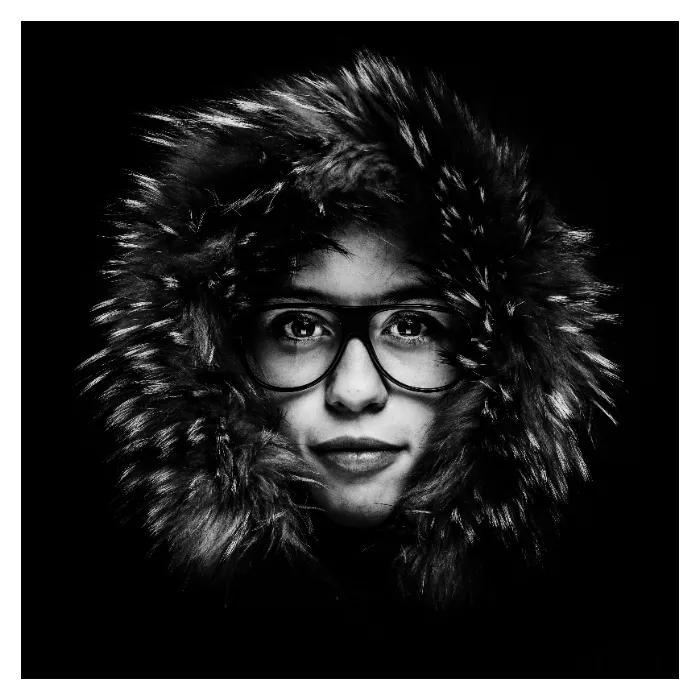 Portrait frontal en noir et blanc d'une femme portant des lunettes et une large capuche en fourrure, contraste marqué sur fond noir, studio Bagnolet.