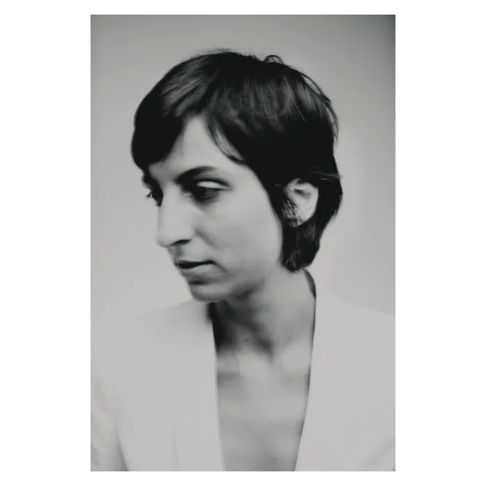 Portrait de trois-quart en noir et blanc d'une femme aux cheveux courts (coupe pixie) regardant vers le bas, portant une veste de costume blanche épurée, photographie de studio à la lumière douce réalisée à Paris par Nicolas Louis.