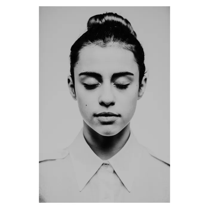 Portrait de face en noir et blanc d'une jeune femme aux yeux clos, visage serein avec un chignon haut, portant une chemise blanche à col pointu, photographie de studio minimaliste réalisée au Vésinet par Nicolas Louis.