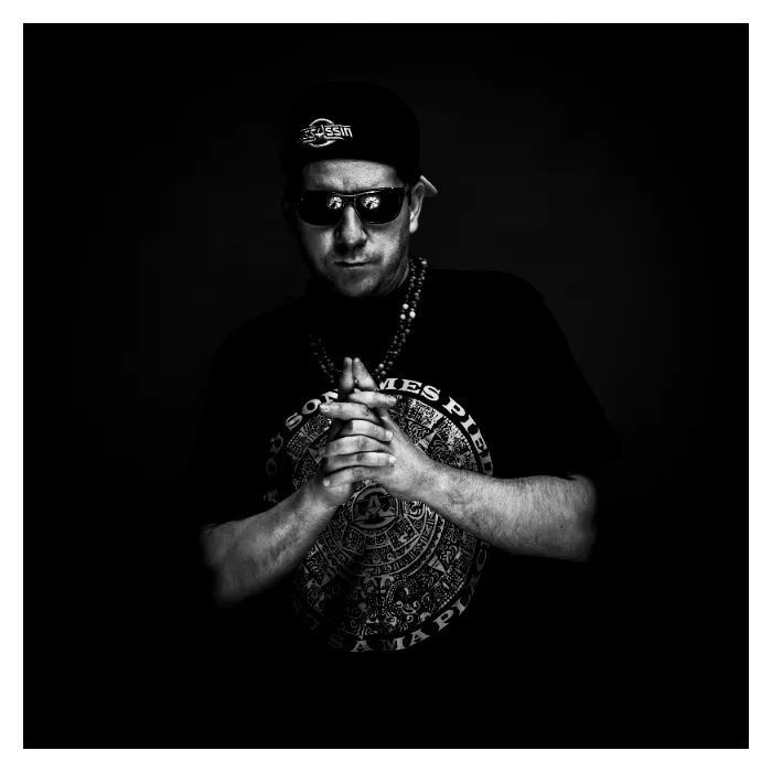 Portrait en noir et blanc de Rockin' Squat (Assassin) en studio, portant une casquette, des lunettes de soleil et un t-shirt graphique avec un collier de perles, mains jointes dans une posture charismatique sur fond sombre, photographie réalisée à Aubervilliers par Nicolas Louis.