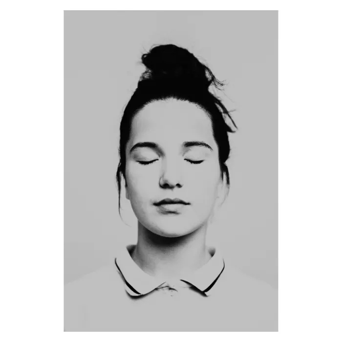 Portrait de face en noir et blanc d'une jeune femme au regard frontal et paisible, visage encadré par un chignon haut et quelques mèches folles, portant un polo blanc à liseré sombre, photographie de studio épurée réalisée au Vésinet par Nicolas Louis.