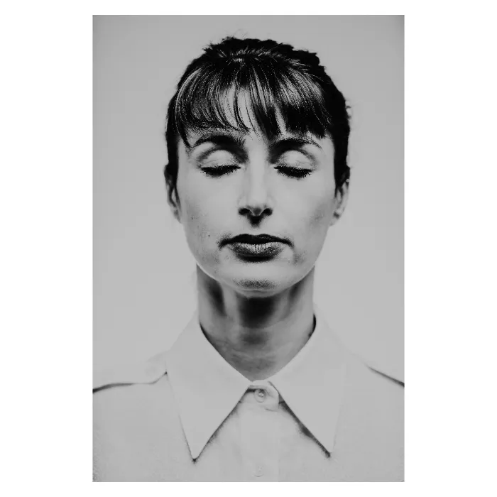 Portrait de face en noir et blanc d'une femme aux yeux clos, visage serein avec une frange droite et des cheveux attachés, portant une chemise blanche boutonnée, photographie de studio minimaliste réalisée au Vésinet par Nicolas Louis.