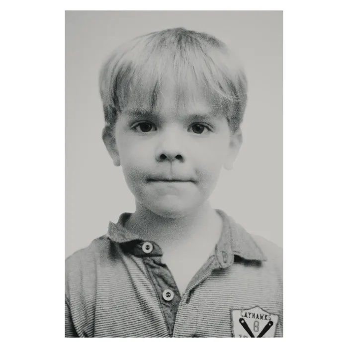 Portrait de face en noir et blanc d'un jeune garçon au regard calme, cheveux blonds avec une frange, portant un polo rayé avec un écusson 'CatHawks', photographie de studio épurée réalisée à Aubervilliers par Nicolas Louis.