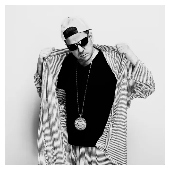 Portrait en noir et blanc de Rockin' Squat (Assassin) debout sur fond blanc, les mains écartant un long gilet rayé sur un t-shirt noir, portant une casquette blanche et des lunettes de soleil, photographie de studio à l'esthétique hip-hop réalisée à Aubervilliers par Nicolas Louis.