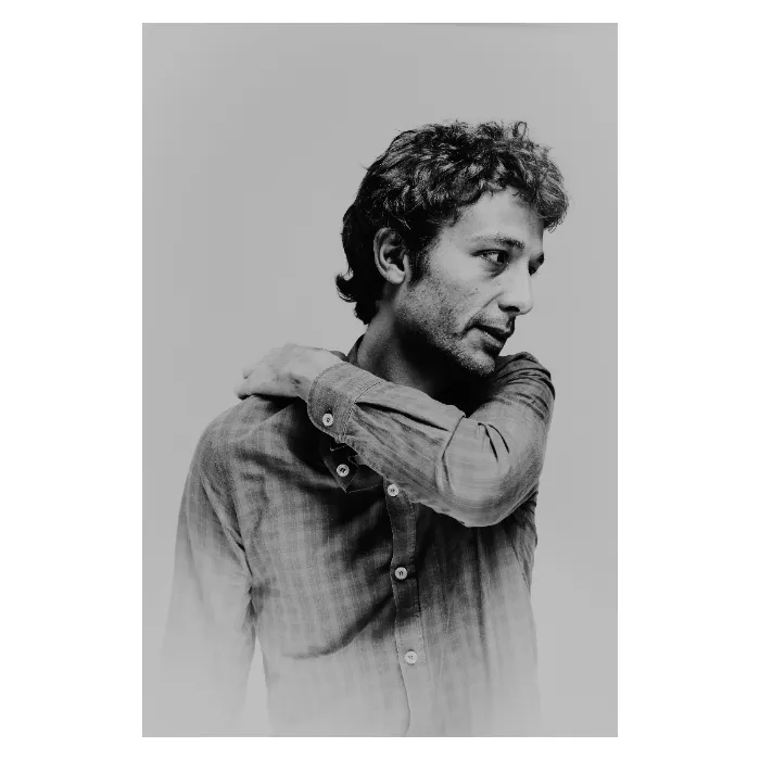 Portrait de profil en noir et blanc d'un homme aux cheveux bouclés, la main posée sur l'épaule opposée dans une pose pensive, portant une chemise à carreaux, photographie de studio épurée réalisée à Bagnolet par Nicolas Louis.