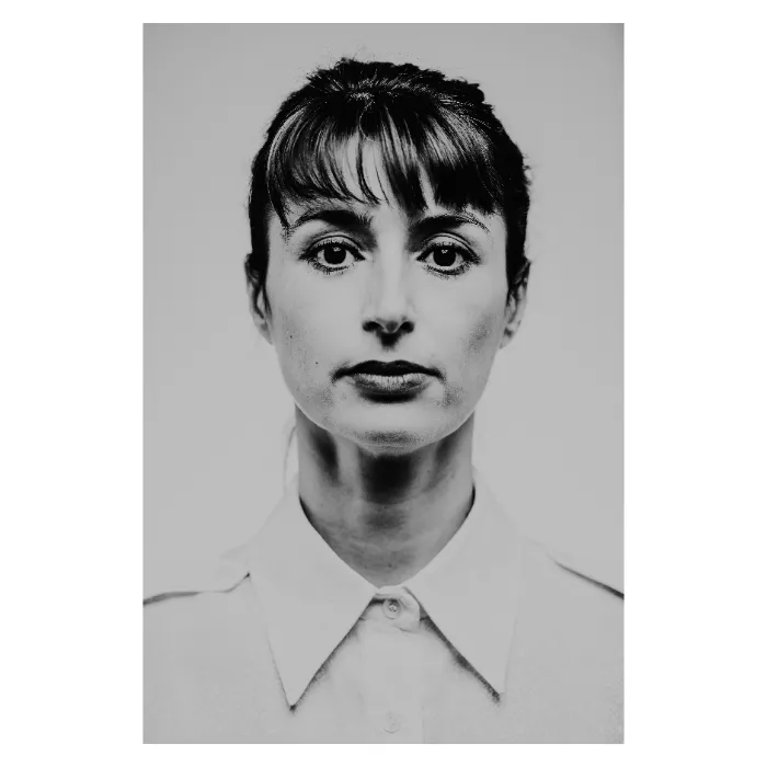 Portrait de face en noir et blanc d'une femme au regard direct et soutenu, visage encadré par une frange droite et des cheveux attachés, portant une chemise blanche boutonnée, esthétique de studio épurée réalisée au Vésinet par Nicolas Louis.