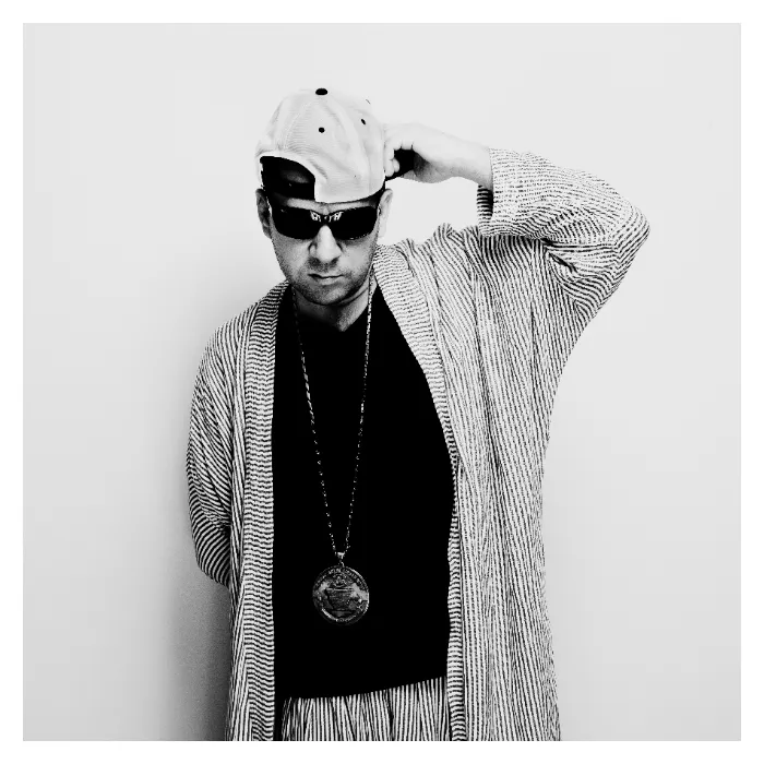 Portrait en noir et blanc de Rockin' Squat (Assassin) sur fond blanc, ajustant une casquette blanche portée à l'envers, lunettes de soleil noires et long gilet rayé, esthétique urbaine épurée, photographie de studio réalisée à Aubervilliers par Nicolas Louis.