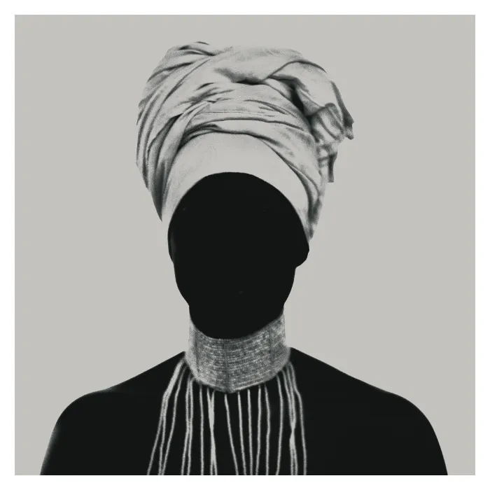 Photographie d'art en noir et blanc montrant une silhouette féminine en contre-jour total, visage masqué par l'ombre et surmonté d'un turban imposant, portant un collier de perles tombantes, studio Le Vésinet par Nicolas Louis.