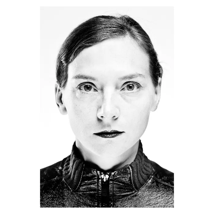 Portrait frontal en noir et blanc d'une femme au regard fixe et déterminé, éclairage contrasté mettant en valeur le grain de la peau, portant une veste technique zippée à col montant, photographie de studio réalisée à Bagnolet par Nicolas Louis.