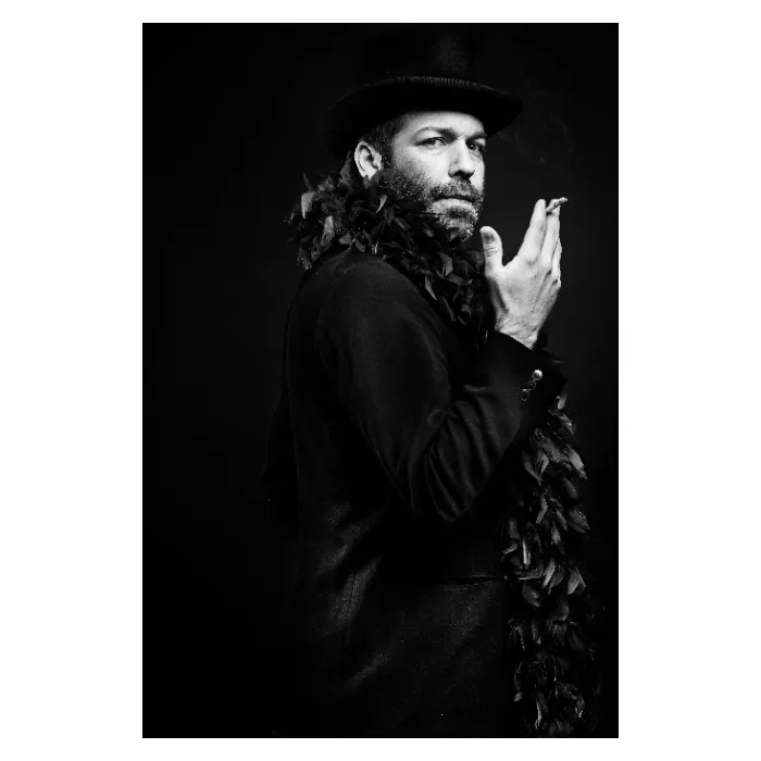 Portrait de profil en noir et blanc d'un homme barbu portant un chapeau haut-de-forme, un boa en plumes et tenant une cigarette allumée, regardant vers l'objectif sur un fond noir profond, photographie de studio à l'ambiance cinématographique réalisée à Bagnolet par Nicolas Louis