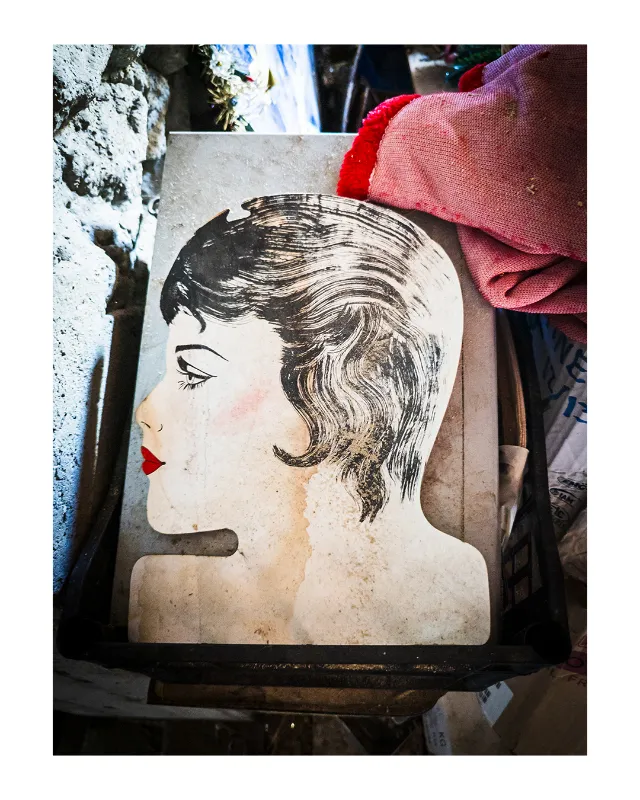 Portrait stylisé d'une femme aux cheveux ondulés années 1920, lèvres rouges, sur affiche vintage patinée exposée parmi tissus colorés dans une brocante à Dieulefit, Drôme