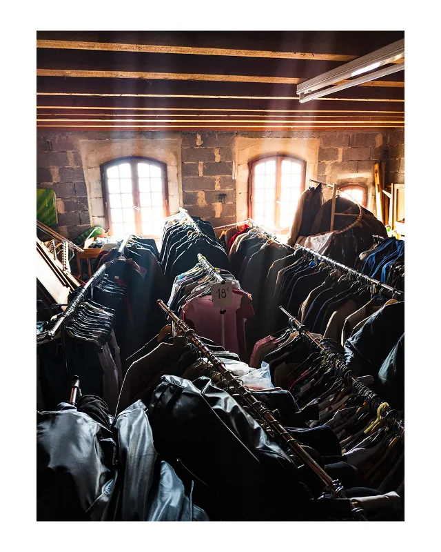 Vestiaire vintage des Grands Magasins Universels à Dieulefit, rangées denses de vêtements noirs sur portants, lumière naturelle dorée traversant trois fenêtres cintrées en pierre, poutres apparentes au plafond