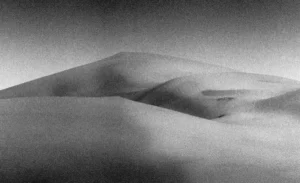 Photographie noir et blanc granuleuse de dunes du désert au Maroc, aux courbes sensuelles et abstraites évoquant des formes corporelles, baignées d'une lumière diffuse et d'une atmosphère onirique, style écriture photographique minimaliste, par le photographe nicolas louis.