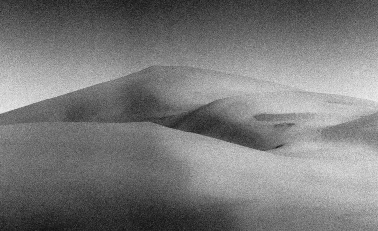 Photographie noir et blanc granuleuse de dunes du désert au Maroc, aux courbes sensuelles et abstraites évoquant des formes corporelles, baignées d'une lumière diffuse et d'une atmosphère onirique, style écriture photographique minimaliste, par le photographe nicolas louis.