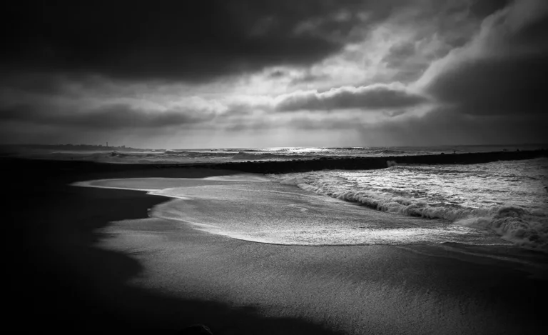 Photographie noir et blanc d'un paysage marin à Anglet : vagues écumantes sur sable mouillé, ciel chargé de nuages dramatiques laissant filtrer une lumière rasante sur l'horizon, avec la silhouette de Biarritz en fond. Longue pose, ambiance tempétueuse.