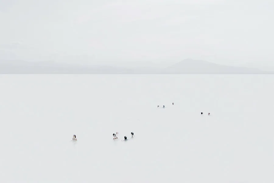 Photographie de la série photo Mar Corpus, en high-key et plan très large : petits groupes de nageurs alignés dans une mer laiteuse, horizon net et reliefs montagneux diffus au loin, composition minimaliste centrée sur l’espace et la profondeur esthétique, réalisée à Biarritz, par Nicolas Louis