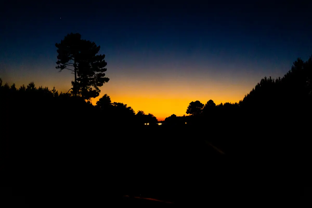 photographie de reportage, d'un paysage d'autoroute en Gironde au couché de soleil, dans un dégradé du bleu au orange, avec la silhouette des arbres.