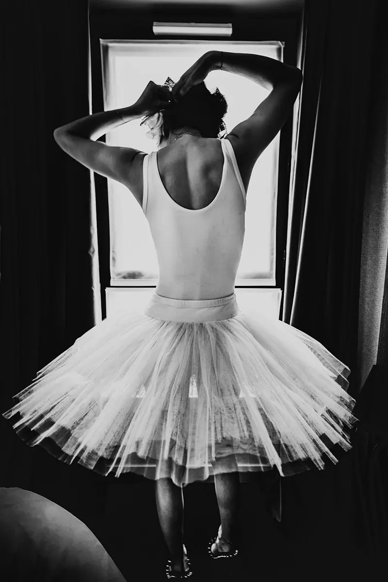 Le modèle féminin Lala, de dos devant la fenêtre, juste au corps et tutu en clair-obscur, se raccrochant les cheveux. Série de portrait intime " Afternoon with Lala" dans un hotel, par le photographe Nicolas Louis à Paris.