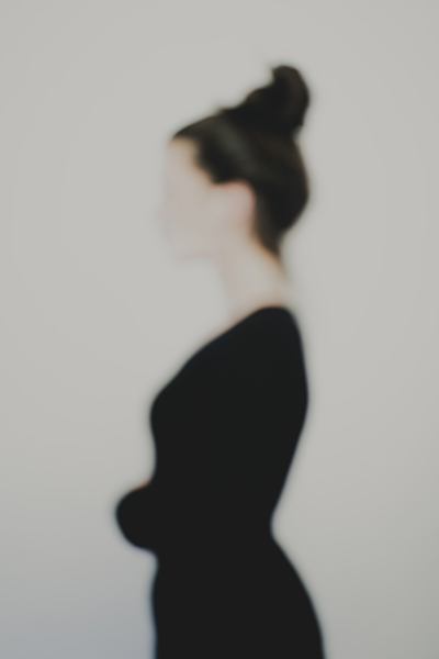 Photographie artistique défocalisée : silhouette féminine de profil, chignon et tenue sombre, contours adoucis et identité estompée, fond neutre minimaliste — extrait de la série de portrait « Erasure », par le photographe Nicolas Louis, réalisé au Vésinet, en France.