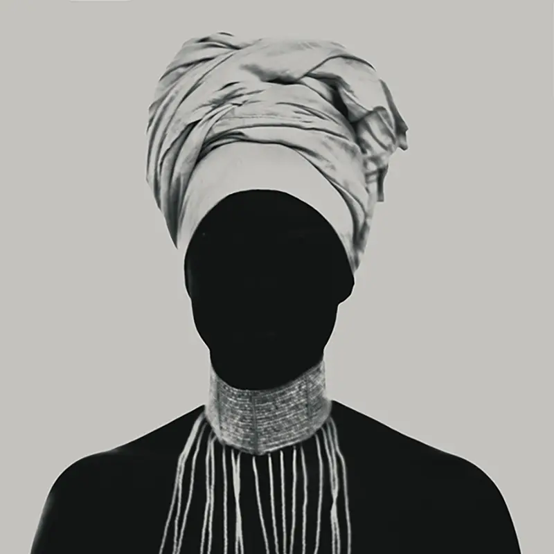 Photographie d'art en noir et blanc montrant la chanteuse fFafa Ruffino, en contre-jour total, visage masqué par l'ombre et surmonté d'un turban imposant, portant un collier de perles tombantes, studio photo, Le Vésinet par Nicolas Louis en 2016.