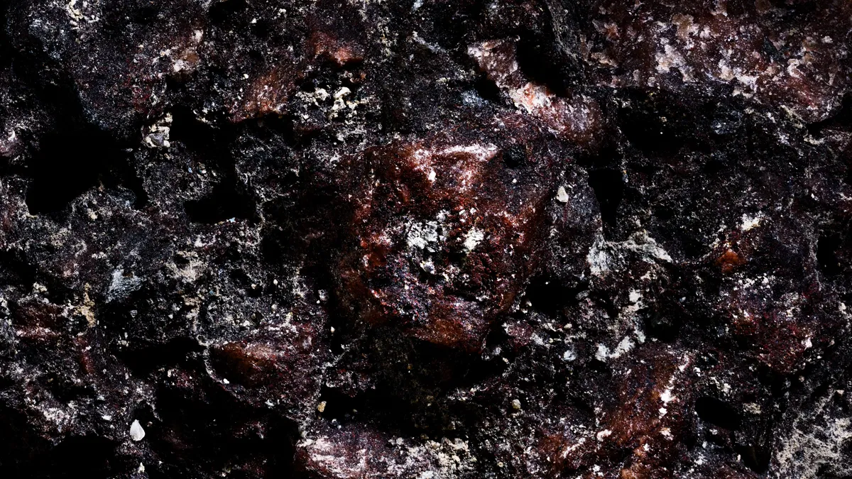 Macro-photographie d'art en studio montrant le gros plan d'une roche volcanique très sombre et rugueuse, caractérisée par des cavités profondes, des textures magmatiques d'un rouge-bordeaux violacé et de légers éclats cristallins clairs.