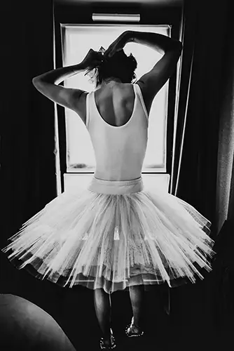 Le modèle féminin Lala, de dos devant la fenêtre, juste au corps et tutu en clair-obscur, se raccrochant les cheveux. Série de portrait intime " Afternoon with Lala" dans un hotel, par le photographe Nicolas Louis à Paris.