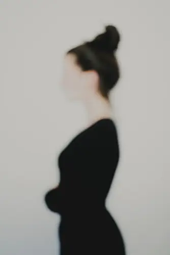 Photographie artistique défocalisée : silhouette féminine de profil, chignon et tenue sombre, contours adoucis et identité estompée, fond neutre minimaliste — extrait de la série de portrait « Erasure », par le photographe Nicolas Louis, réalisé au Vésinet, en France.