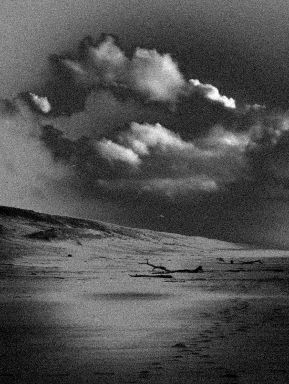 Photographie artistique en noir et blanc d’un paysage cotier désert, avec ciel nuageux et plage balayee par le vent. Style granuleux et minimaliste, evoquant la solitude, le silence et la poesie du littoral.